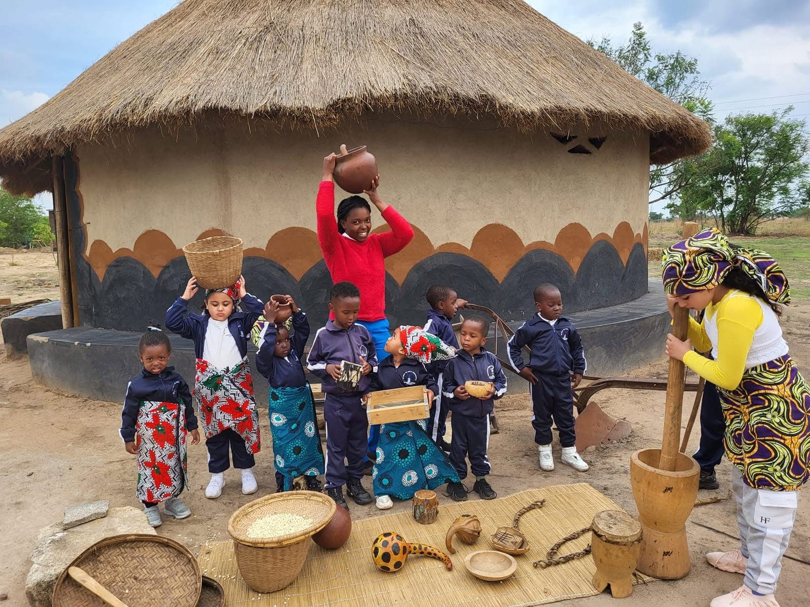Children carrying baskets and pots in front of a thatched hut on cultural day at Nirmeen